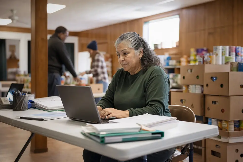 A food pantry volunteer treasurer working on a laptop beside stacked boxes of canned goods.