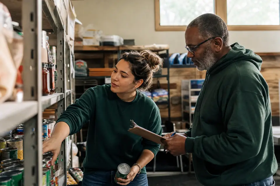Two food pantry volunteers sorting donated cans on shelves in a church basement.