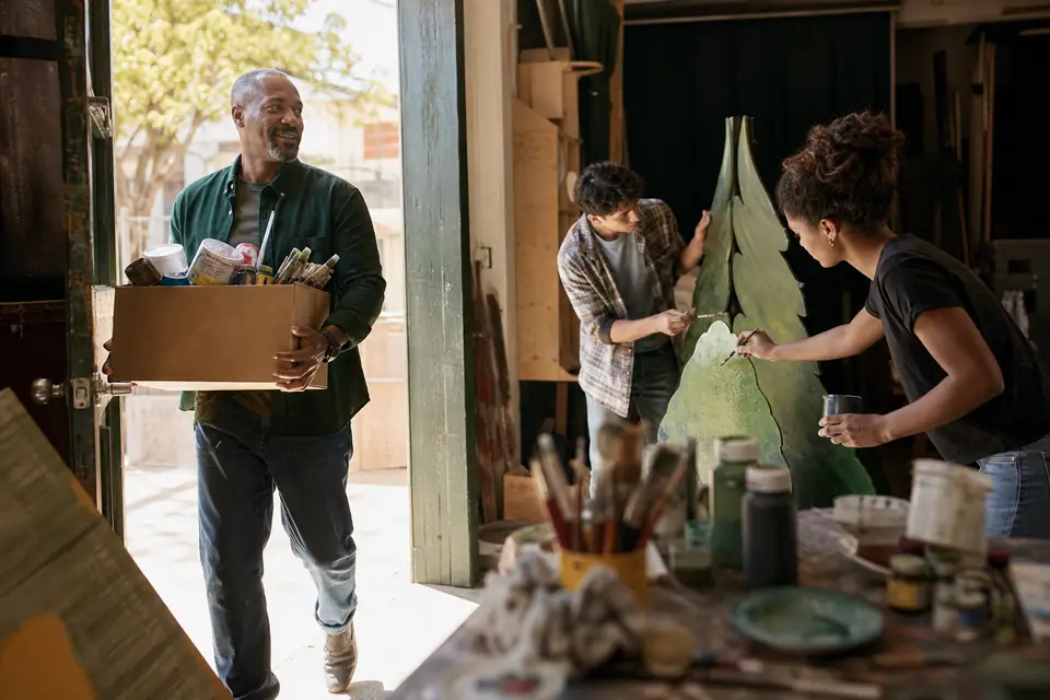 Theater director carrying donated supplies into a small arts studio with teen volunteers.